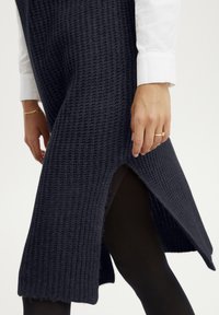 Strickkleid in Marineblau mit strukturiertem Muster, seitlichem Schlitz und sichtbarem Saum. Das Model trägt schwarze Leggings mit einem weißen Hemd unter dem Kleid.