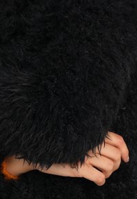 Sort, pelslignende overtøj med en struktureret overflade. Har en løs pasform og brede manchetter, hvor en hånd hviler på stoffet.