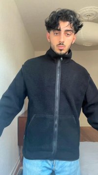 Black fleece zip-up jacket with high collar and side pockets, worn over blue jeans. Soft texture with a relaxed fit.