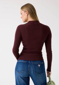 Burgundy bordázott kötött felső magas nyakkal, testhezálló dizájnnal, párosítva kék farmerral, amelynek hátsó zsebében logó díszítés található.