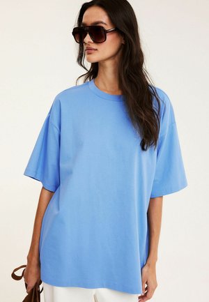 Vrouw die een oversized lichtblauw T-shirt en grote donkere zonnebril draagt, een bruine handtas vasthoudend, tegen een effen witte achtergrond.