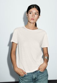 Hvid t-shirt med korte ærmer i en afslappet pasform, lavet af blødt stof, parret med blå jeans. Modellen bærer et sølv cuff-armbånd.