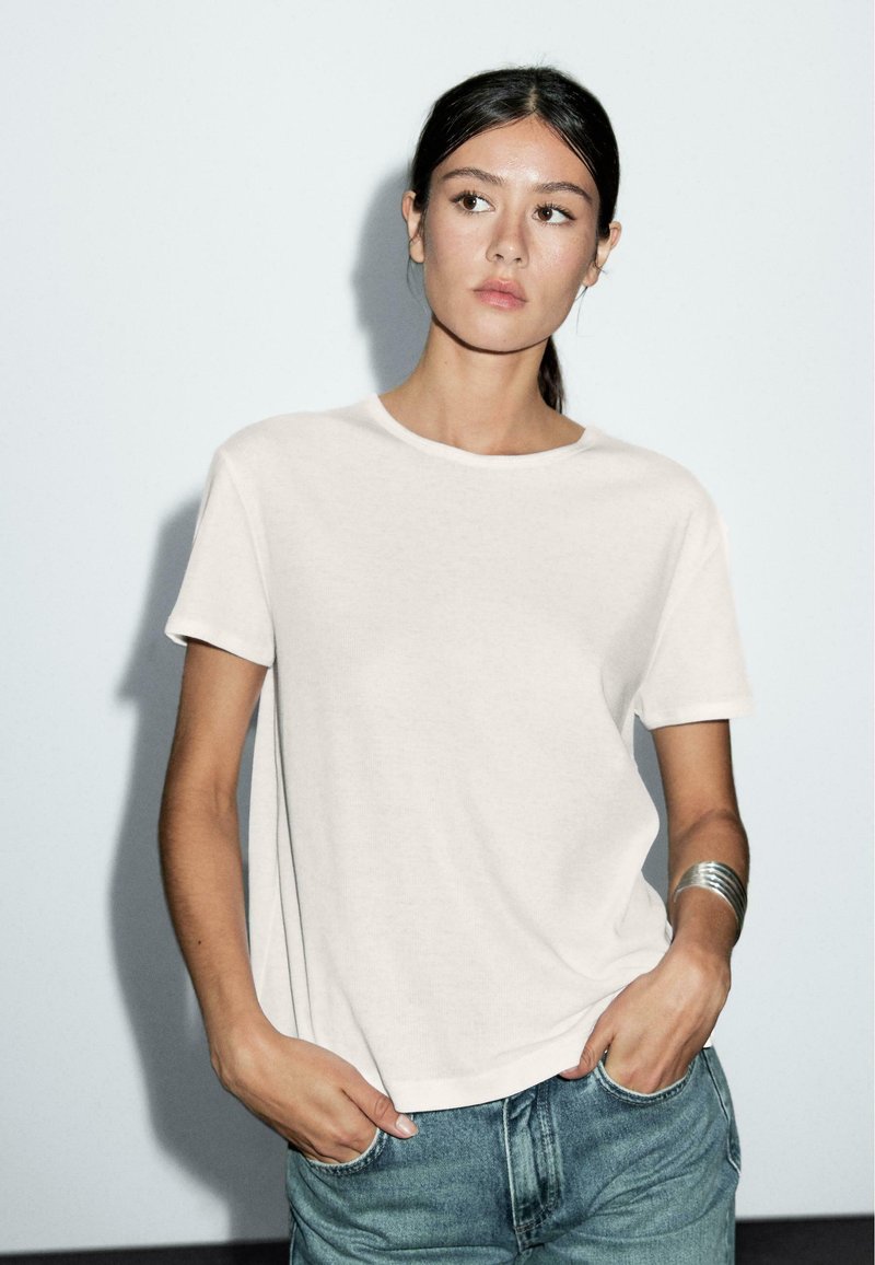 Hvid t-shirt med korte ærmer i en afslappet pasform, lavet af blødt stof, parret med blå jeans. Modellen bærer et sølv cuff-armbånd.