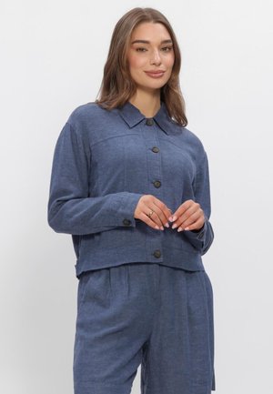 Frau trägt ein blaues, langärmeliges Hemd mit Knöpfen und passende weite Hose, steht mit leicht verschränkten Händen vor neutralem Hintergrund.