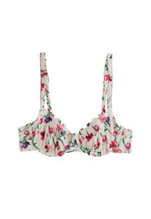 Femme portant un bikini avec un motif floral et des bords festonnés, debout devant un mur en pierre avec des fleurs rouges à proximité.