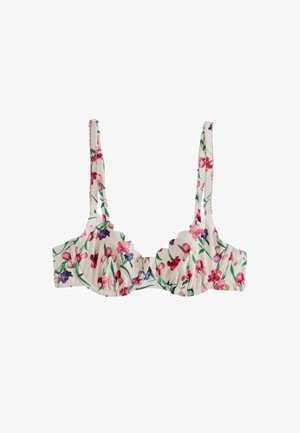 Kobieta w bikini w kwiatowy wzór z falistymi brzegami stoi przed kamiennym murem z czerwonymi kwiatami w pobliżu.