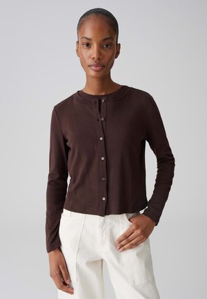 Donna che indossa un cardigan marrone scuro a maniche lunghe con bottoni e pantaloni a vita alta color avorio, in piedi davanti a uno sfondo chiaro e semplice.