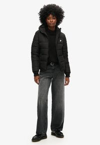 Doudoune noire à capuche, équipée d'une fermeture éclair et d'un logo griffé. Portée par-dessus un pull en maille noire et associée à un jean gris à jambes larges.