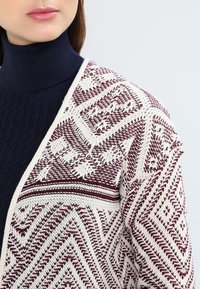 Cardigan lavorato a maglia in bordeaux e crema, con motivi geometrici e tessuto testurizzato. Indossato sopra un dolcevita blu navy per un contrasto.