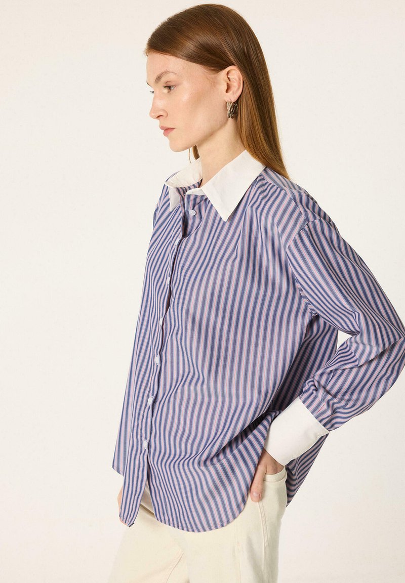 Femme aux cheveux roux lisses portant une chemise rayée bleue et rose à boutons avec col et poignets blancs, posant avec une main dans la poche.