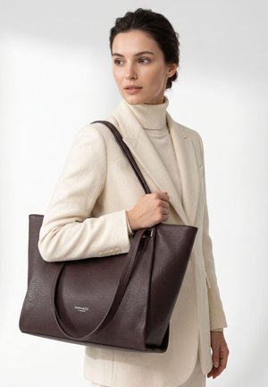 Frau in cremefarbenem Blazer und Rollkragenpullover hält große dunkelbraune Ledertasche mit Schulterriemen, steht vor weißem Hintergrund.