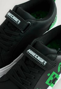 Zapatillas sintéticas negras con gráficos verdes con patrón de Minecraft, que cuentan con un cuello acolchado, cordones y una correa de velcro con logo.