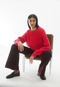 Rode geribbelde gebreide trui met lange mouwen, gecombineerd met wijde bordeauxrode ribfluwelen broek en zwarte slip-ons, zittend op een stoel.