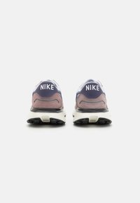 Zapatillas Nike con parte superior en púrpura y azul marino, logo blanco, texturas de gamuza y malla, y suela de goma negra. La vista trasera muestra los detalles del diseño.