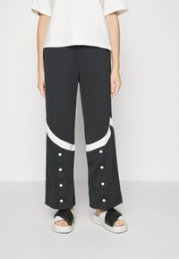 Person som bär svarta vida byxor med vita böjda ränder och tryckknappar vid anklarna, kombinerat med svarta och vita slip-on sandaler.