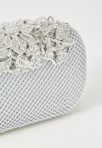 Bolso de mano plateado con malla texturizada y pequeñas cuentas metálicas, adornado con un diseño floral plateado embellecido con rhinestones.