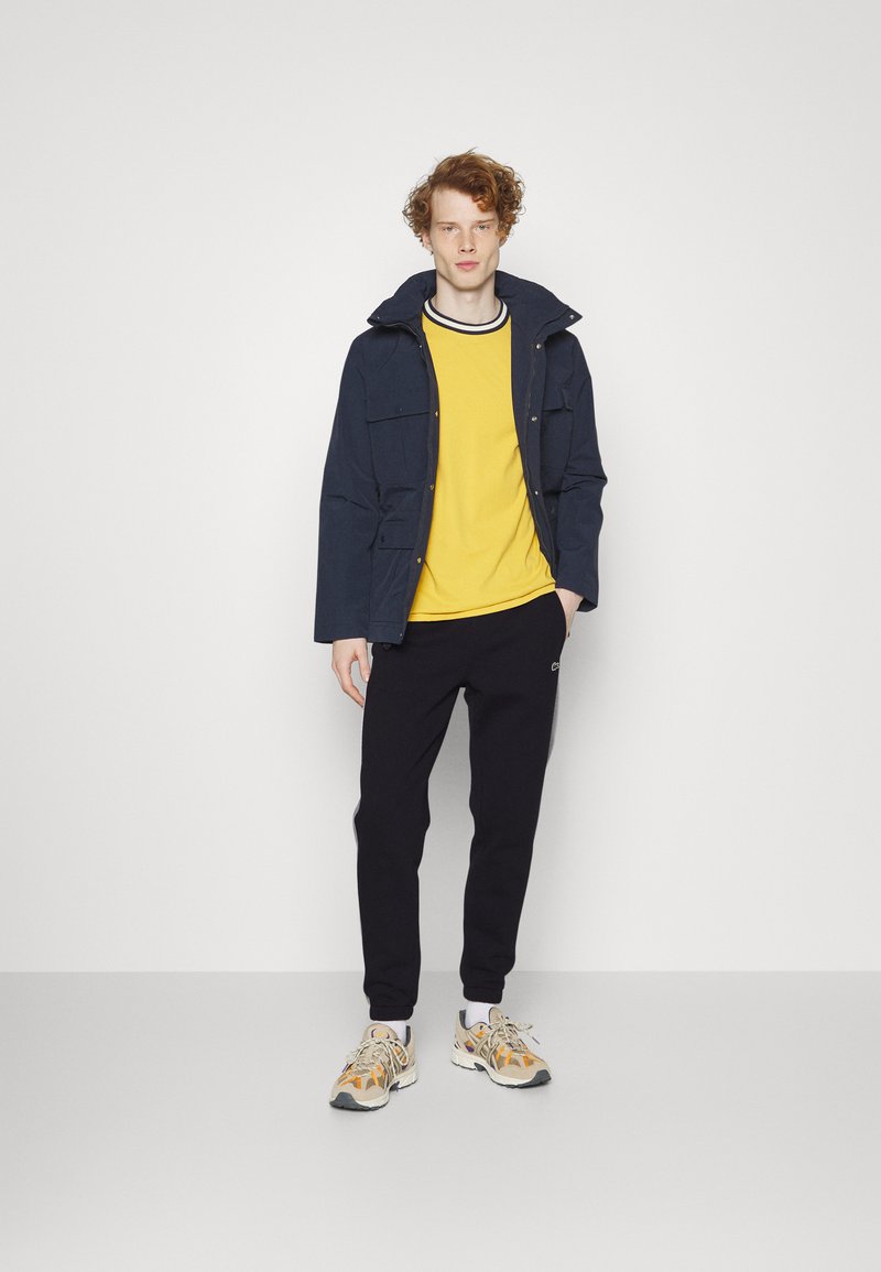 Veste marine, T-shirt jaune avec col noir, pantalon de survêtement noir et baskets à motifs avec accents orange. Design simple et fonctionnel.