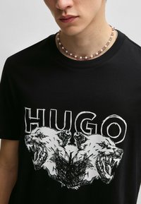 Czarny bawełniany t-shirt z wyrazistym graficznym wzorem przedstawiającym dwie głowy niedźwiedzi oraz słowo "HUGO" w białym kolorze. Dodatkowym akcentem jest okrągły dekolt.
