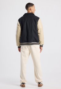 Black wool and beige leather varsity jacket with ribbed cuffs and hem, worn over white trousers and black sneakers. Plain background.
