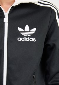 Svart Adidas träningsjacka med vit logga och ränder, dragkedja fram, texturerad tyg, har en sidoficka med dragkedja.