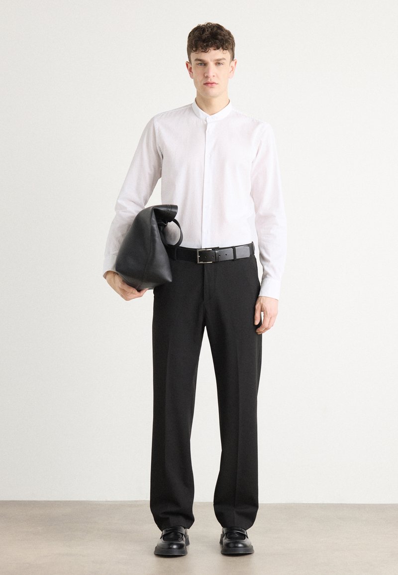 Biała koszula z długim rękawem i kołnierzem mandaryńskim, czarne spodnie, teksturowana czarna torba i czarne buty; model stoi wyprostowany na jednolitym tle.