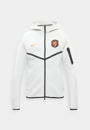 Bijela dukserica s patentnim zatvaračem i crnim obrubom, kapuljačom, narančastim Nike logotipom na prsima i narančastim grbom lava na lijevoj strani.