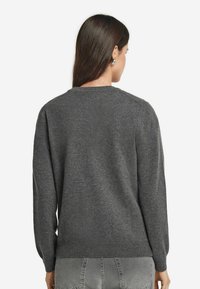 Kobieta z długimi ciemnymi włosami, nosząca srebrne kolczyki w kształcie kół, ciemnoszary sweter i jasnoszare dżinsy, widziana od tyłu na białym tle.