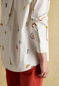 White shirt featuring colorful embroidered patterns of food items and landmarks, with buttoned cuffs and a curved hem, paired with red trousers.