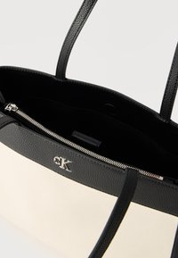 Μονόχρωμη ασπρόμαυρη τσάντα ταχυδρόμου Calvin Klein με διπλές λαβές, ορατό εσωτερικό διαμέρισμα με φερμουάρ και ασημένιο λογότυπο "cK" στην εξωτερική μπροστινή πλευρά.