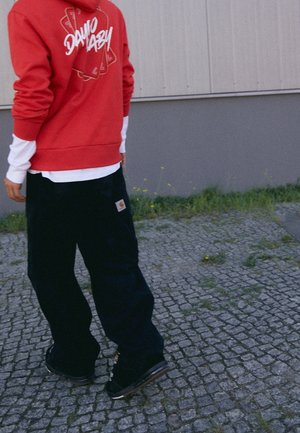 Osoba w czerwonej bluzie z kapturem z motywem kart do gry, warstwowa biała koszula, czarne luźne spodnie i czarne sneakersy na kostiumowych płytach.