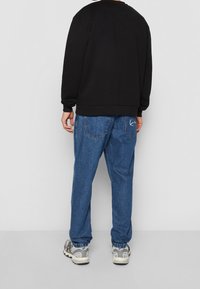 Sudadera negra con cuello redondo, combinada con unos vaqueros de denim azul holgados que tienen bolsillos traseros y bordado de logo en blanco. Zapatillas grises.