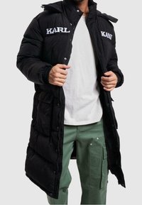 Svart puffjacka med dragkedja och tryckknappsstängning, med "KARL"-logotyp i vitt, sidofickor och huva. Bärs över en vit skjorta.