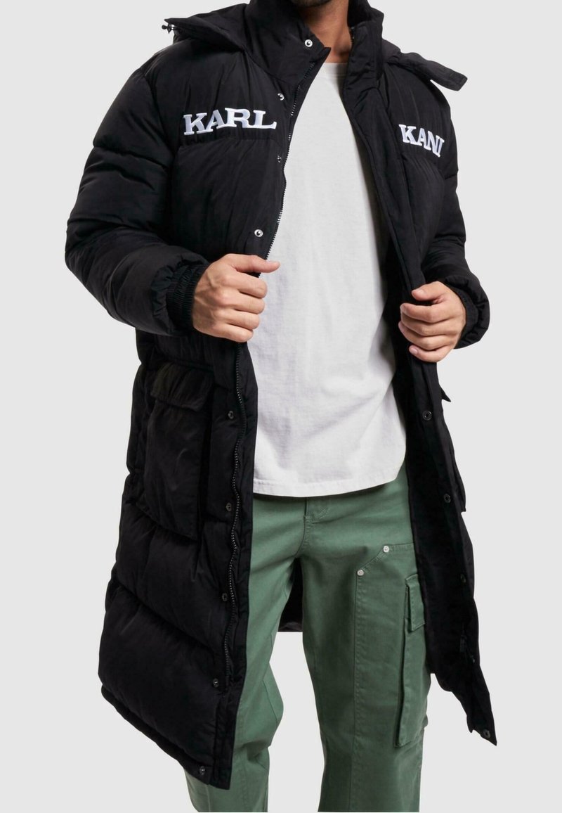 Svart puffjacka med dragkedja och tryckknappsstängning, med "KARL"-logotyp i vitt, sidofickor och huva. Bärs över en vit skjorta.