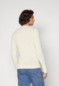 Light beige long-sleeve sweater in a soft, knit texture with a round neckline. Features ribbed cuffs and waistband, worn over blue jeans.