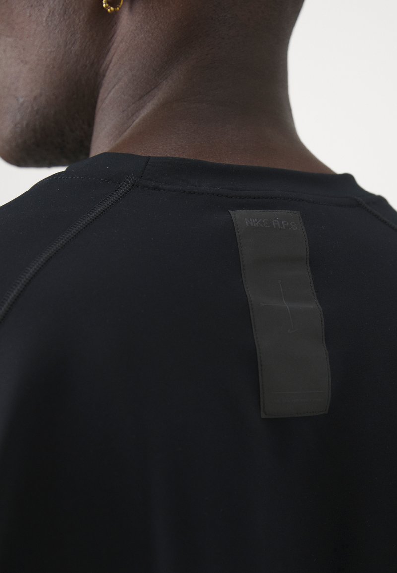 Camiseta atlética negra con una textura suave, que presenta una etiqueta negra cosida en la parte posterior con "NIKE A.P.S." grabado en el centro.