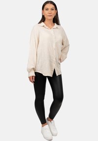 Camicia button-up beige chiaro con un motivo testurizzato, abbinata a leggings neri lucidi e sneakers bianche. La camicia ha un colletto e una vestibilità ampia.