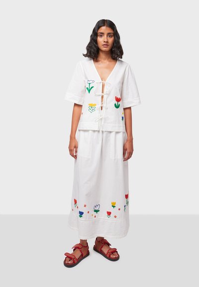 Witte katoenen outfit bestaande uit een top met korte mouwen en een vetersluiting aan de voorkant, versierd met bloemembroidery, gecombineerd met een lange rok en rode sandalen.