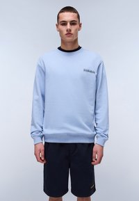 Lys blå sweatshirt i myk stoff, med rund hals og ribbestrikkede mansjetter, med en liten logo på venstre bryst. Brukt med mørke shorts.
