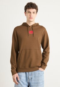 Brun hettegenser med en rød logo-patch, snor i hetten og frontlomme; myk tekstur med ribbede mansjetter og nederkant; avslappet passform.