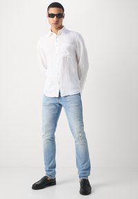 Hvit lin skjorte med knappestolpe og brystlomme, lyseblå denimjeans med slitte detaljer, og svarte loafers.