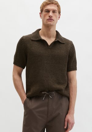 Férfi, aki sötétbarna rövid ujjú kötött galléros pólót és hozzáillő barna zsinóros nadrágot visel, kezét zsebre téve áll.