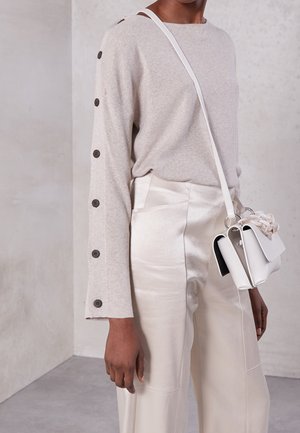 Persona vistiendo una blusa beige de manga larga abotonada, pantalones de cintura alta color crema brillante y llevando un pequeño bolso blanco cruzado con detalle de cadena.