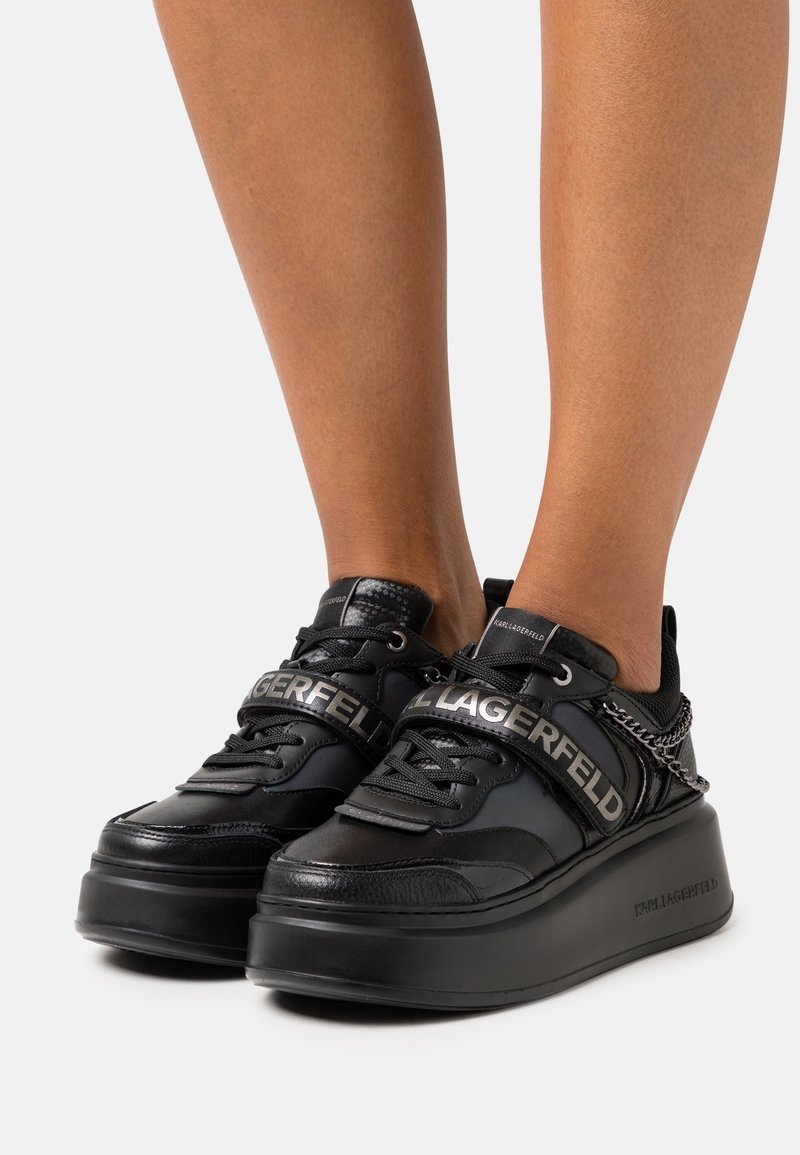 Svarta sneakers med en kraftig plattform, med en texturerad läderovandel, snörning och ett framträdande logoband över fotryggen.