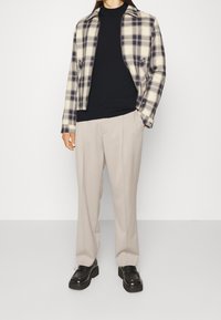 Veste à carreaux crème et marine, t-shirt noir, pantalon beige clair et chaussures noires à semelle épaisse. Coupe droite avec manches longues.