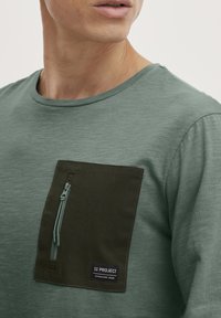 Grünes kurzärmliges Hemd mit strukturiertem Stoff. Es hat eine dunkelgrüne Brusttasche mit Reißverschluss und ein aufgenähtes Markenlabel.