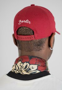 Cască de baseball roșie cu "Berlin" brodat în alb, asociată cu un fular cu model floral în roșu, crem și negru.