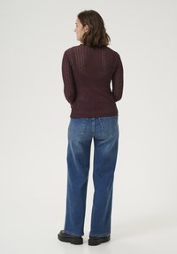 Haut long à manches en maille bordeaux avec un tricot aéré, torse ajusté et motif texturé, associé à un jean en denim bleu taille haute et des bottes noires.