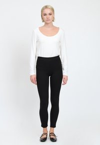 Witte lange mouwen top met een ronde hals, gecombineerd met zwarte leggings met een hoge taille en zijknopen, gedragen met zwarte schoenen.