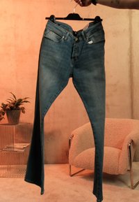 Roka tur turētāju ar asimetriskām zilām džinsu biksēm telpā ar bēšu krēslu un vadu sānu galdiņu ar augu un žurnāliem.