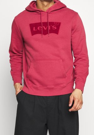 Man wearing a red Levi's hoodie with front pocket and drawstrings, paired with black pants, standing against a plain background.